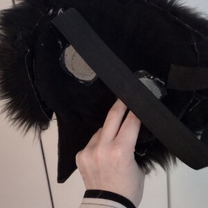 May include: A black furry mask with an elastic strap, likely for a costume or cosplay. The mask has a textured, plush appearance and features mesh eye coverings. The person holding the mask is wearing a black and white striped shirt.