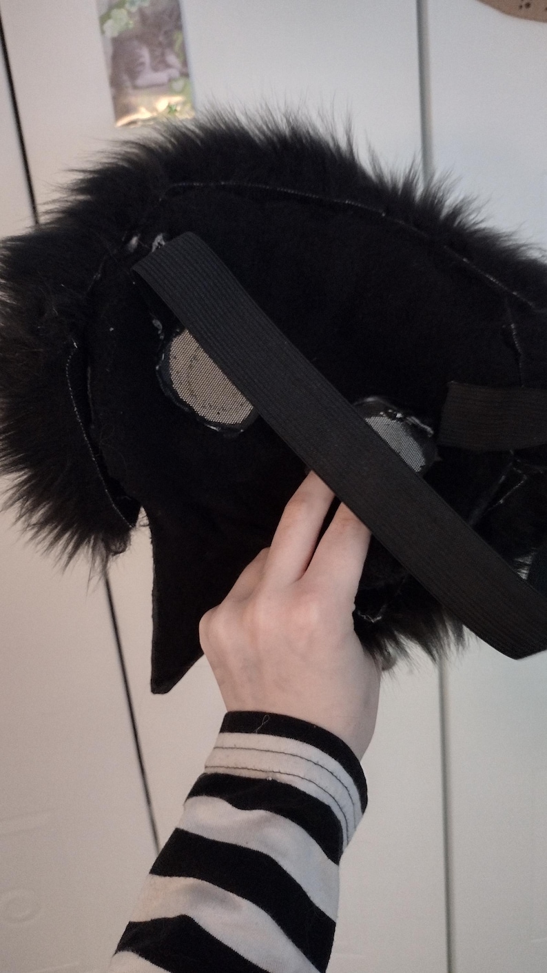 May include: A black furry mask with an elastic strap, likely for a costume or cosplay. The mask has a textured, plush appearance and features mesh eye coverings. The person holding the mask is wearing a black and white striped shirt.