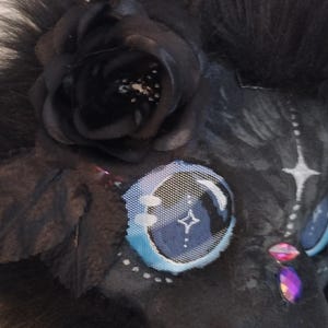 May include: Close-up of a black furry mask with a black rose and blue and white eye details. The mask has a star design on the eye and is decorated with pink and purple gems.