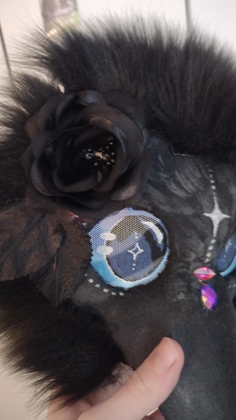 May include: Close-up of a black furry mask with a black rose and blue and white eye details. The mask has a star design on the eye and is decorated with pink and purple gems.