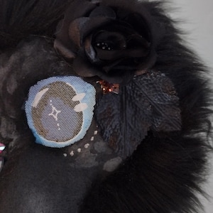May include: A dark-colored, furry mask with a black rose and leaf embellishments. A circular patch with a blue and white design is also attached. The mask has a textured appearance and is likely handmade.