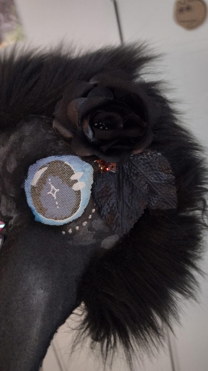 May include: A dark-colored, furry mask with a black rose and leaf embellishments. A circular patch with a blue and white design is also attached. The mask has a textured appearance and is likely handmade.