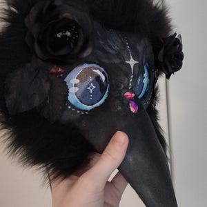 May include: A black plague doctor mask with a long beak, adorned with black faux fur, black roses, and jewel accents. The eyes are detailed with blue and white patterns. A unique costume accessory.