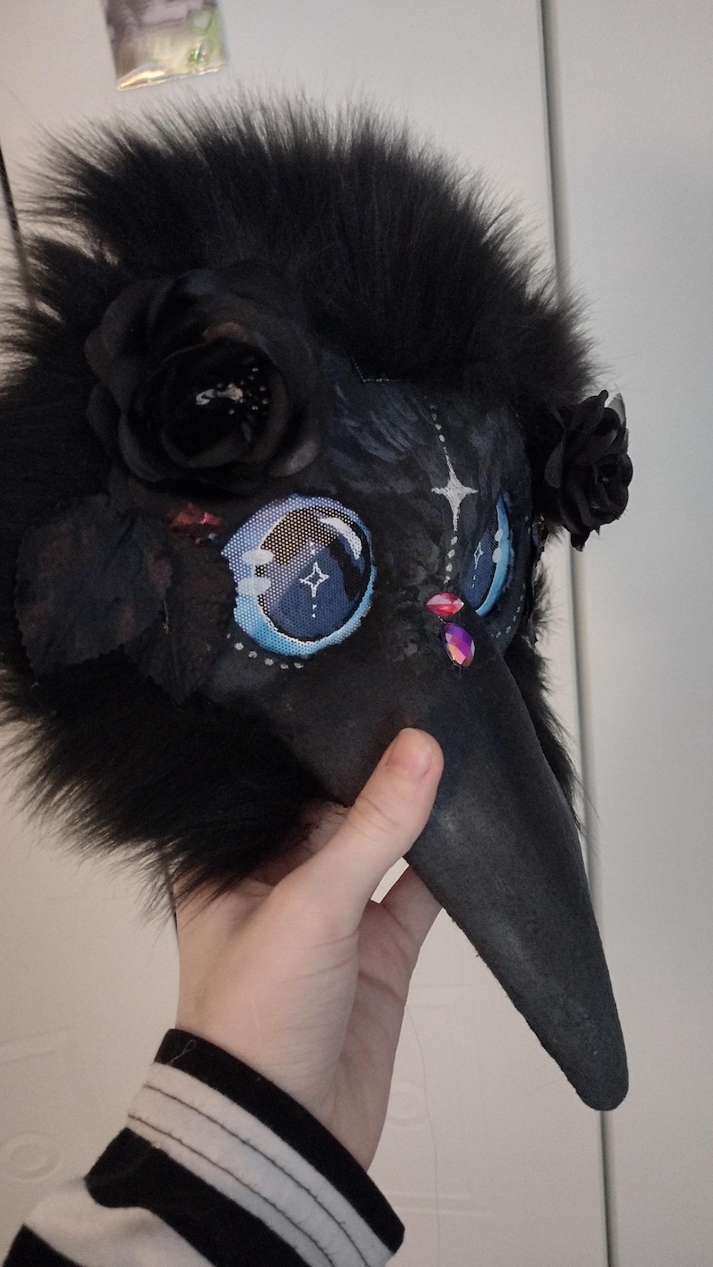 May include: A black plague doctor mask with a long beak, adorned with black faux fur, black roses, and jewel accents. The eyes are detailed with blue and white patterns. A unique costume accessory.