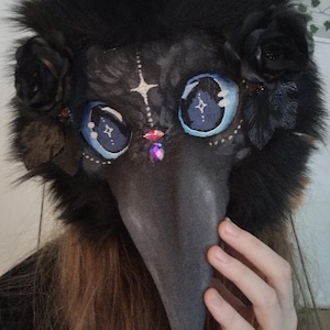 May include: A black plague doctor mask with a long, gray beak and large, blue eyes with star-like designs. The mask is adorned with black roses and a fluffy, black fur trim. A hand is visible on the right side of the mask.