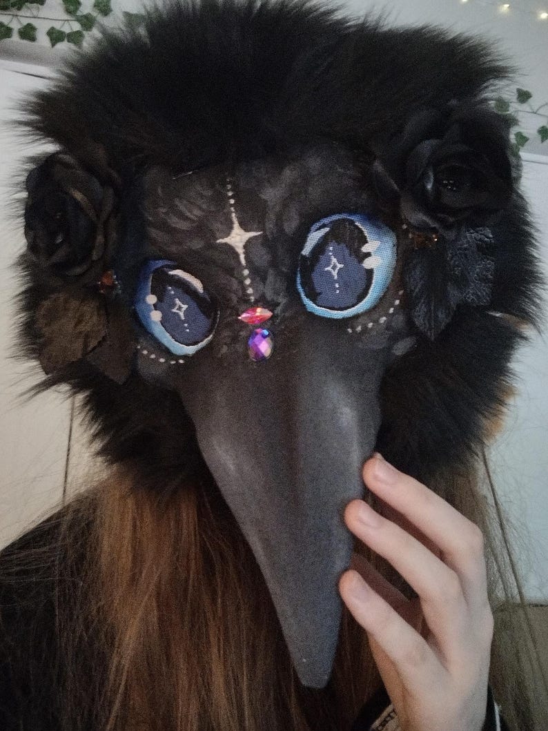 May include: A black plague doctor mask with a long, gray beak and large, blue eyes with star-like designs. The mask is adorned with black roses and a fluffy, black fur trim. A hand is visible on the right side of the mask.