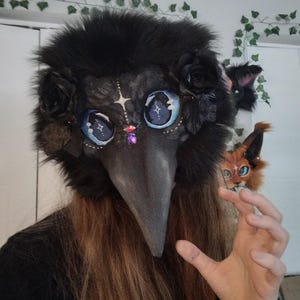 May include: A black plague doctor mask with a long beak, adorned with black roses and fluffy fur. The mask features large, detailed blue eyes with star-like designs and a purple gem. A small, orange and white fox-like creature is also visible.