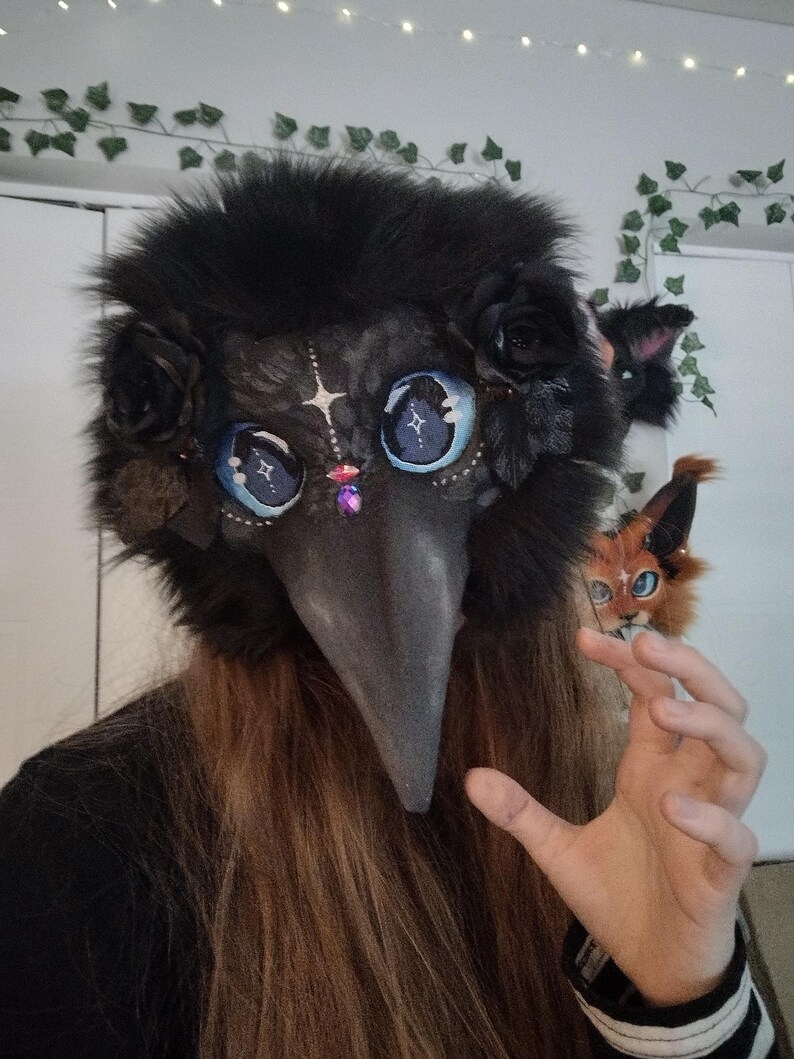 May include: A black plague doctor mask with a long beak, adorned with black roses and fluffy fur. The mask features large, detailed blue eyes with star-like designs and a purple gem. A small, orange and white fox-like creature is also visible.
