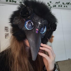 May include: A black plague doctor mask with a long, gray beak. The mask features large, blue eyes with star details, black roses, and a fluffy black fur trim. Decorative jewels are placed around the eyes. The mask is designed for cosplay or costume use.