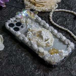 May include: A white iPhone case decorated with white and gold 3D frosting, pearls, and iridescent butterflies. The case features a small angel figurine in a white frame.