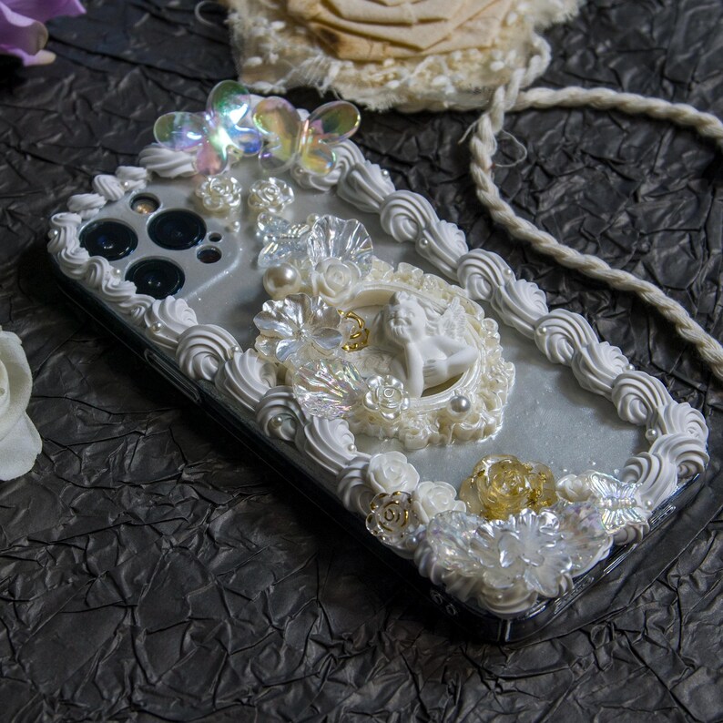 May include: A white iPhone case decorated with white and gold 3D frosting, pearls, and iridescent butterflies. The case features a small angel figurine in a white frame.