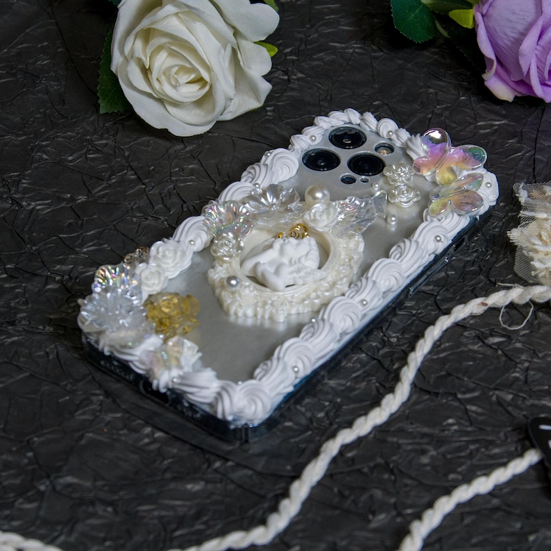 May include: A white phone case with a decorative design featuring white frosting, pearls, and a white angel figurine. The case has a silver background and a black camera lens.