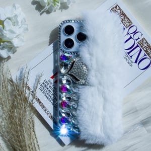 Puede incluir: Una funda de teléfono de piel sintética blanca con la parte posterior transparente y un diseño de diamantes de imitación. La funda tiene un gran diseño de flor de diamantes de imitación y diamantes de imitación más pequeños alrededor del objetivo de la cámara.