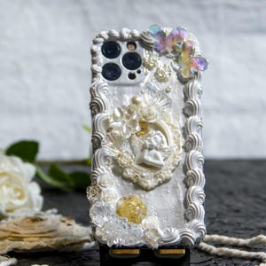 May include: A white phone case decorated with white and gold 3D embellishments, including flowers, a cherub, and a butterfly. The case has a white background with a pearl-like finish.