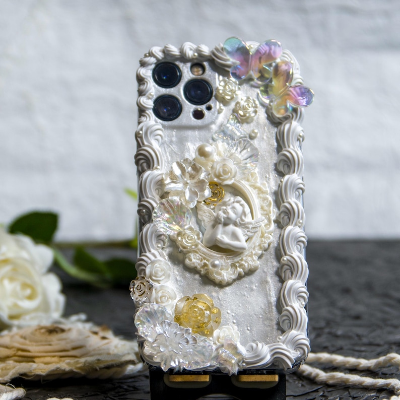 May include: A white phone case decorated with white and gold 3D embellishments, including flowers, a cherub, and a butterfly. The case has a white background with a pearl-like finish.