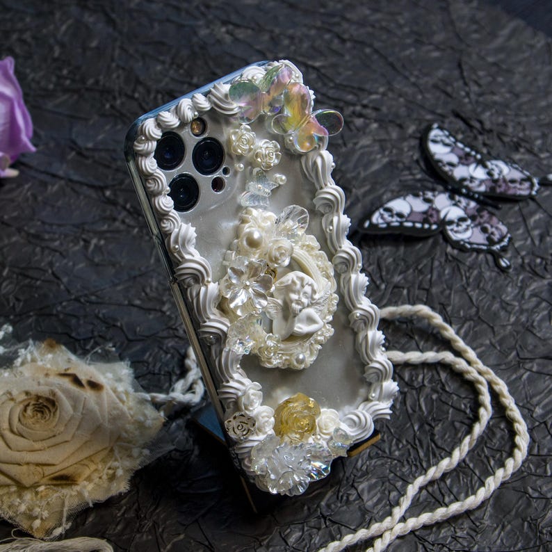 May include: A white phone case with a decorative design featuring white frosting, pearls, and a small angel figurine. The case has a white rope strap attached.