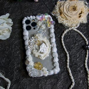 May include: White iPhone case with a decorative design featuring a cherub, flowers, and pearls. The case has a white, fluffy border and a white strap.