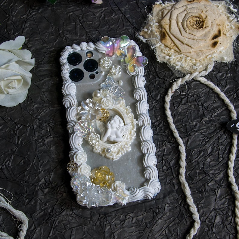 May include: White iPhone case with a decorative design featuring a cherub, flowers, and pearls. The case has a white, fluffy border and a white strap.