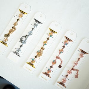 May include: Five decorative watch bands with floral and heart accents. The bands are in gold, silver, and rose gold tones, each with a different design. The watch bands are displayed on white cards.