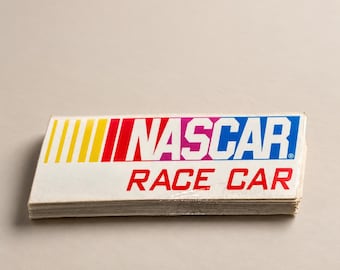 May include: A rectangular sticker featuring the NASCAR logo in blue, red, and yellow, with the text RACE CAR printed in bold red capital letters below. The sticker has a white background and a glossy finish, showing a stack of multiple units.
