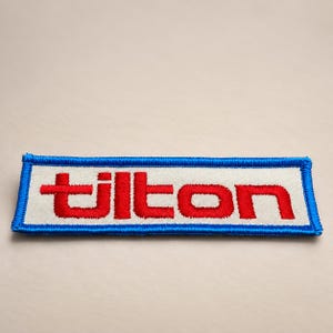 May include: A rectangular embroidered patch featuring a white background with the word tilton stitched in bold red lowercase letters. The patch is finished with a thick, vibrant blue stitched border that frames the entire rectangular shape.