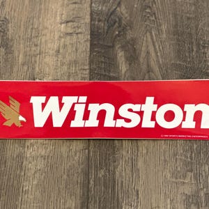Peut inclure: Autocollant rouge et blanc avec le mot "Winston" en lettres blanches et un logo d'aigle doré. L'autocollant porte la mention de copyright "©1983 SPORTS MARKETING ENTERPRISES, INC."