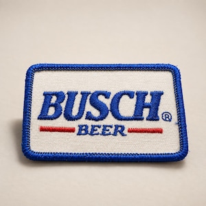 May include: A rectangular embroidered patch with a white background and a blue border. The text BUSCH is embroidered in large blue letters, with the word BEER in smaller blue letters below, flanked by two horizontal red lines. A registered trademark symbol is present.