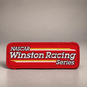 May include: A rectangular red embroidered patch featuring the white text NASCAR Winston Racing Series. The design includes two horizontal yellow and white stripes running across the centre, with a red stitched border surrounding the entire patch.