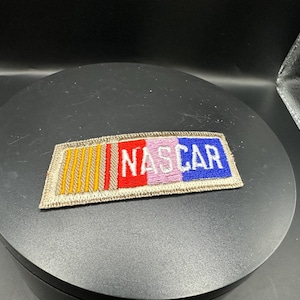Könnte beinhalten: Ein beigefarbener Stoff-Patch mit dem Wort "NASCAR" in weißen Buchstaben gestickt. Der Patch hat ein Design aus roten, rosa und blauen Streifen mit goldenen Streifen auf der linken Seite.