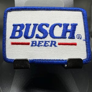 Puede incluir: Un parche blanco bordado con letras azules que dice "Busch Beer" con una franja roja por encima y por debajo del texto. El parche tiene un borde azul.