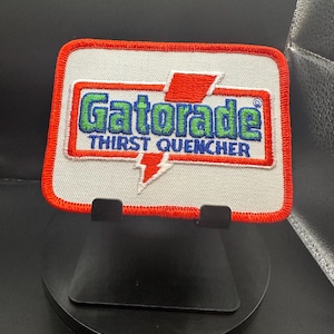 Puede incluir: Un parche blanco bordado con un rayo rojo y las palabras "Gatorade Thirst Quencher" en letras verdes y azules.