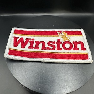 Könnte beinhalten: Ein weißer, roter und goldener gestickter Aufnäher mit dem Wort "Winston" in goldenen Buchstaben und einem stilisierten Adler-Design.