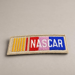May include: A rectangular embroidered patch featuring the text NASCAR in white letters. The design includes vertical stripes in yellow, red, light pink, and blue, all bordered by a tan stitched edge. The patch has a flat, woven texture.