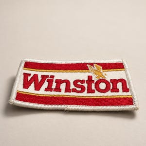 Könnte beinhalten: Ein rechteckiger gestickter Aufnäher mit dem Wort Winston in fetten roten Buchstaben. Das Design umfasst ein weißes Mittelband mit einem goldenen Adleremblem, das oben und unten von roten Streifen begrenzt wird und einen weißen Rand hat.