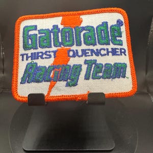 Peut inclure: Un écusson brodé blanc avec une bordure orange. L'écusson représente un éclair et le texte "Gatorade Thirst Quencher Racing Team".