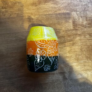 May include: A yellow, orange, and black ceramic vase with a white floral design. The vase has a rounded shape and a wide mouth.