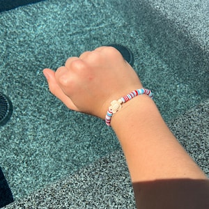 May include: A colorful bracelet with red, white, and blue beads, and a small, light-colored turtle charm. The bracelet is worn on a wrist, with a background of water.