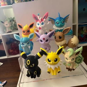 May include: A collection of eight small, colorful plastic figurines representing various fictional creatures. The figures are arranged on a clear tiered display stand, featuring characters with distinct features like large ears, tails, and varied color schemes.