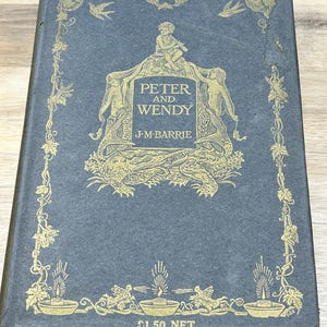 Pode incluir: Livro antigo de capa dura intitulado "Peter and Wendy" de J.M. Barrie. A capa é azul escuro com elementos decorativos dourados, incluindo ilustrações de figuras, folhagens e o título. O livro apresenta desgaste.