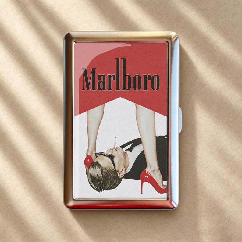 May include: A rectangular metal cigarette case featuring a graphic print of the Marlboro logo in black text over a red background. The image depicts a person in a suit and glasses with a cigarette, framed by legs wearing red high heels.