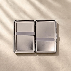 May include: A rectangular silver-toned metal cigarette case shown in an open position. The interior features two smooth, reflective compartments, each equipped with a thin, horizontal metal tension bar designed to hold contents securely in place.