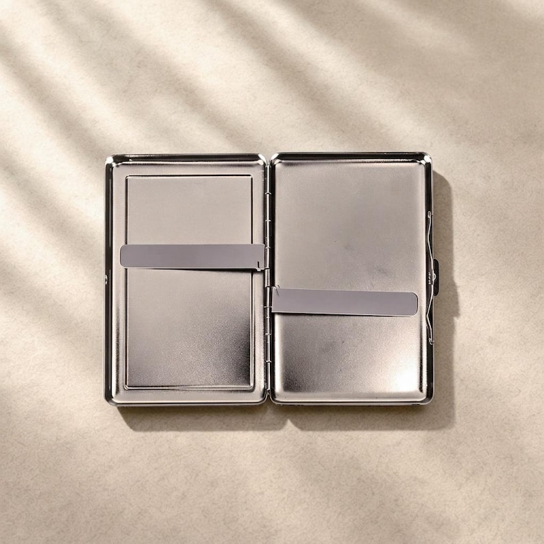 May include: A rectangular silver-toned metal cigarette case shown in an open position. The interior features two smooth, reflective compartments, each equipped with a thin, horizontal metal tension bar designed to hold contents securely in place.