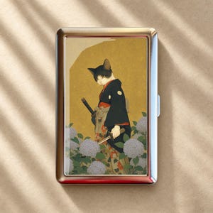 May include: A rectangular metal cigarette case featuring a graphic illustration of a black and white cat dressed in a dark kimono with a sword, standing among light purple hydrangea flowers against a textured gold background.