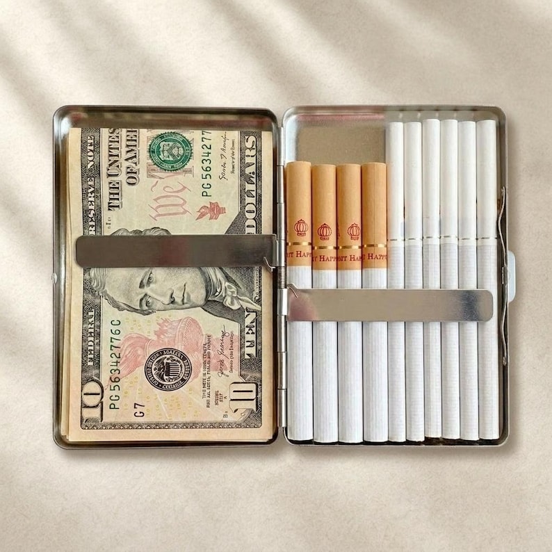 May include: A rectangular metal cigarette case with a hinged opening. The left side holds a ten-dollar bill secured by a silver metal clip. The right side contains nine white cigarettes with tan filters, held in place by a horizontal silver metal bar.