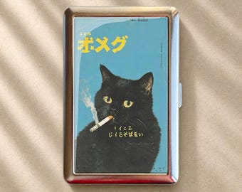 以下が含まれることがあります： 青い背景に、火のついたタバコを口にくわえた黄色い目の黒猫のグラフィックがプリントされた、長方形の銀色の金属製シガレットケース。猫のイラストの上下には黄色い日本語のテキストが配置されています。