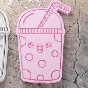May include: Two cookie cutters shaped like a bubble tea drink. One is white, the other is pink. Both have a smiling face, bubbles, and a straw. The cutters are on a wooden surface.