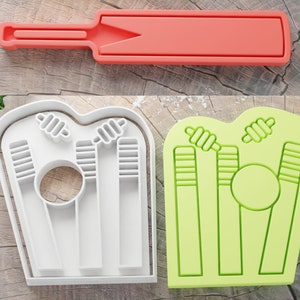 May include: Two white plastic cookie cutters, one shaped like a cricket wicket with three stumps and a ball, and the other shaped like a cricket bat.