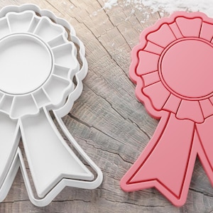 May include: Two plastic cookie cutters shaped like a ribbon with a circle in the center. The cutter on the left is white and the cutter on the right is red.