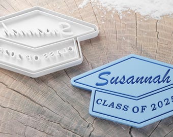 May include: Two graduation cap-shaped cookie cutters. One is white and says "Class of 2025". The other is blue and says "Susannah Class of 2025".