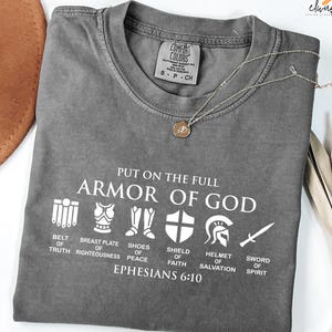 May include: Gray t-shirt with a white graphic of the Armor of God. The graphic includes a belt, breastplate, shoes, shield, helmet, and sword. The text "Put on the full Armor of God" and "Ephesians 6:10" are also included in the graphic.
